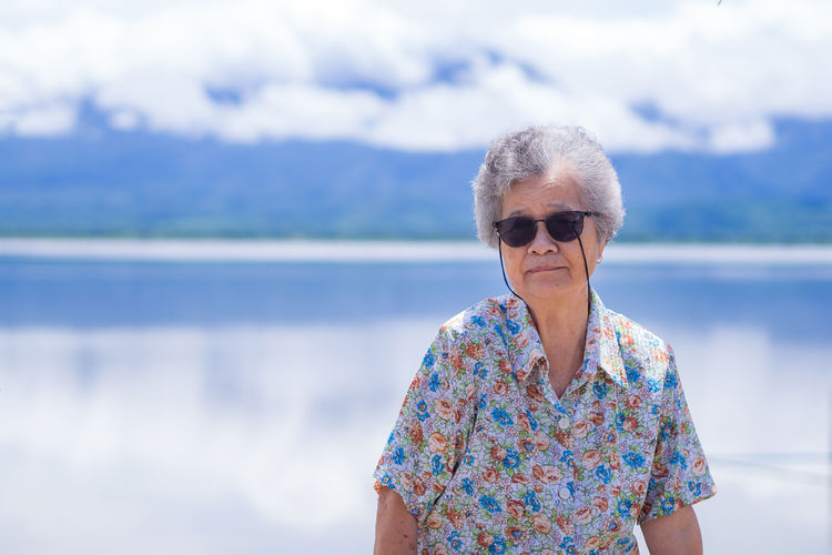 Senior woman wearing sunglasses and sitting on chair side the lake with mountains background.