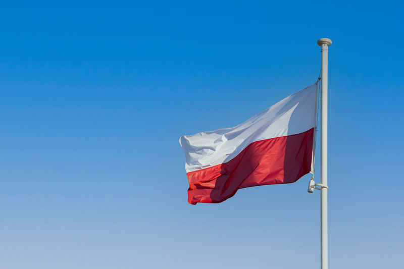 The flag of poland flying in the wind on a tall pole against a blue sky.
