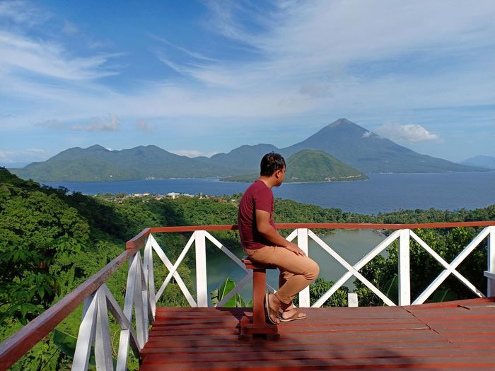 Full length of man sitting at observation point against sea and mountains