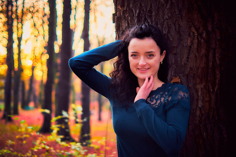 Portrait of smiling woman standing by tree trunk in forest