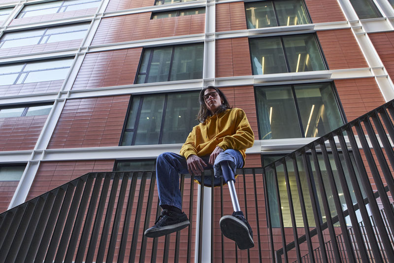 Young man with leg prosthesis sitting on metal railing against building