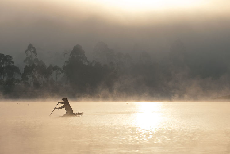 Man sitting in lake against sky during sunset