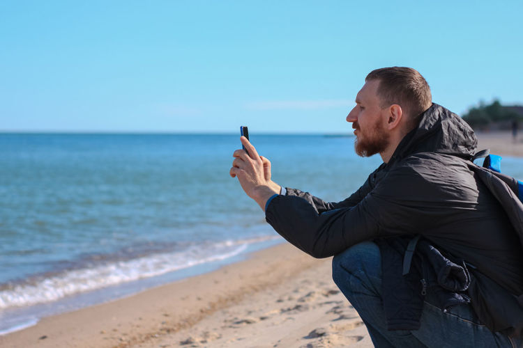 Side view of man using mobile phone at beach against clear sky