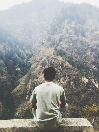 Rear view of man looking at mountains while sitting on concrete wall