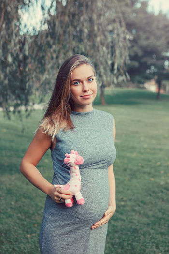 Portrait of smiling pregnant young woman holding stuffed toy by abdomen while standing on grass