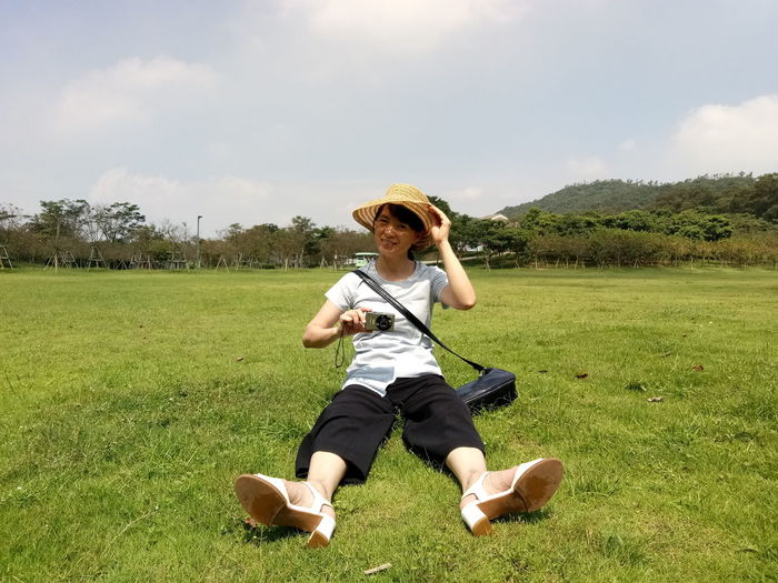 Portrait of woman photographing while sitting on field against sky
