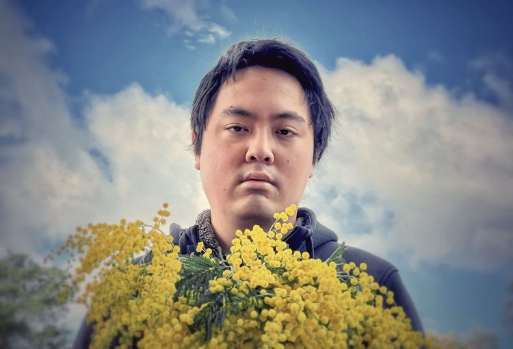 Portrait of young man holding golden wattle flowers against cloudy blue sky.