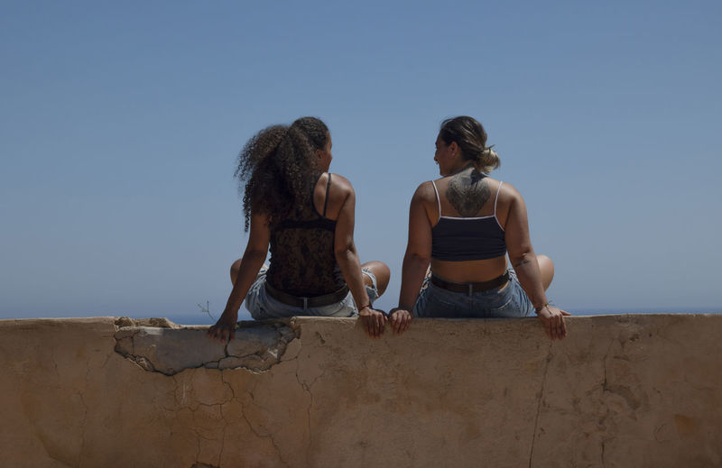 Rear view of 2 women sitting on wall beside the sea against clear blue sky