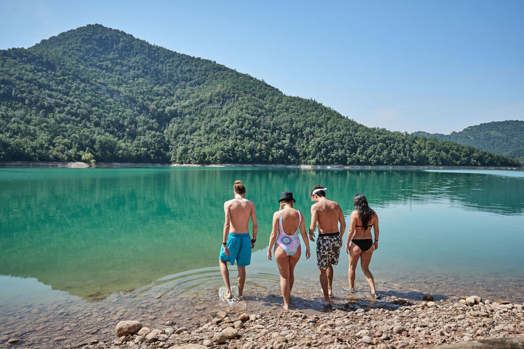 Back view of company of tourists in swimwear walking together in calm lake on sunny day during vacation in highland area