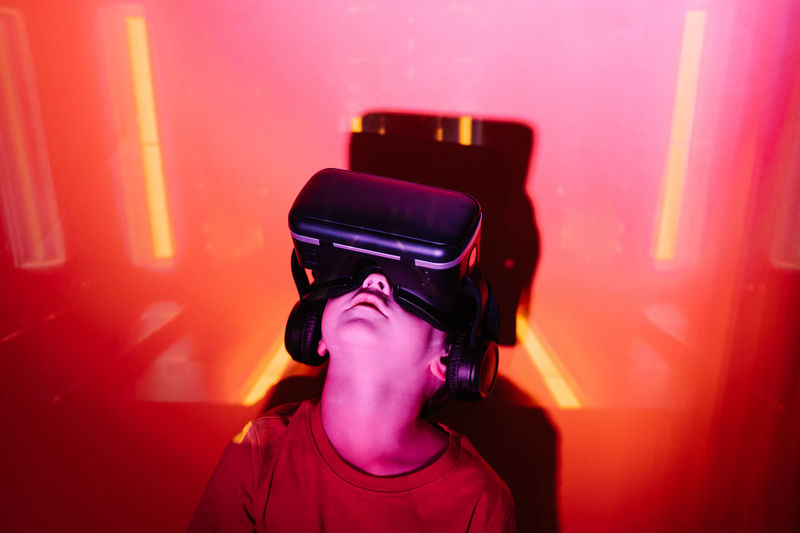 High angle of little boy with raised arms leaning on wall and exploring virtual reality under red neon light