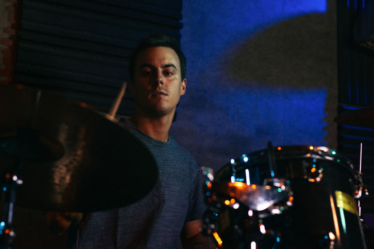 Concentrated young male musician playing drums in club with green and blue neon illumination