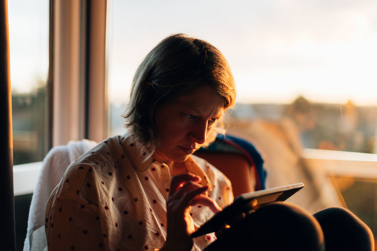 A woman sitting indoors nearby a big window at sunset using a tablet. wireless technology concept