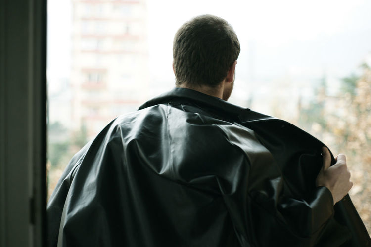 Rear view of man removing black leather jacket by window