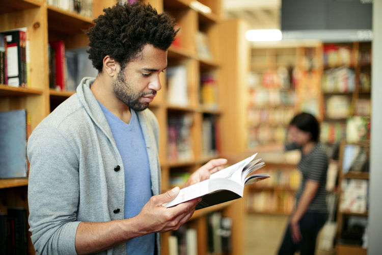 Man reading book in library with woman standing in background
