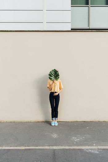 Woman holding monstera leaf in front of face by wall