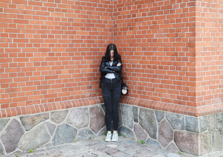 Young beautiful brunette girl in black jacket and jeans stands near wall of gothic church on summer
