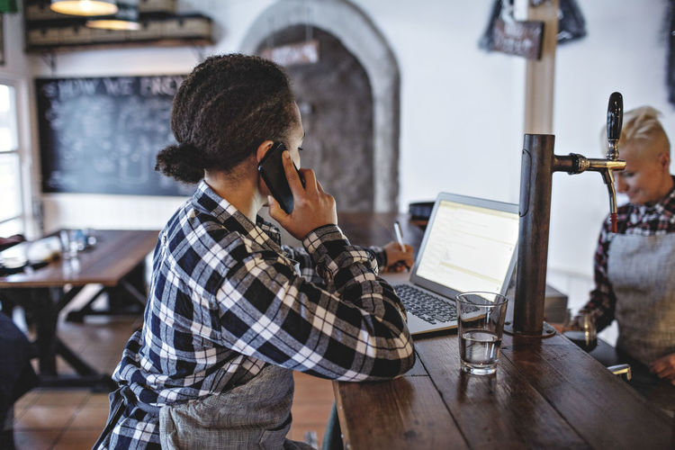 Female manager talking on mobile phone while using laptop at bar counter