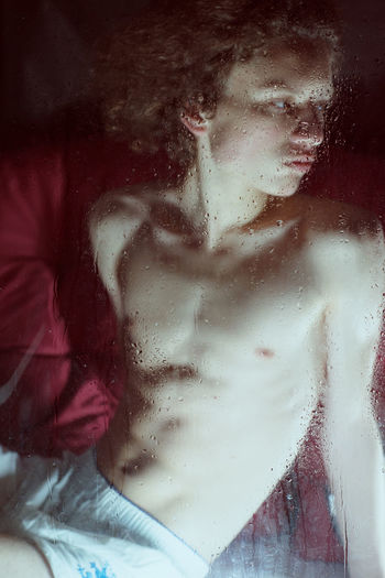 Shirtless man sitting at home seen through wet window