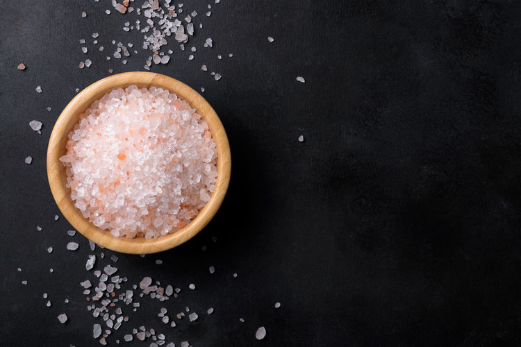 Pink himalayan salt in a wooden bowl with scattered salt on a black background, copy space