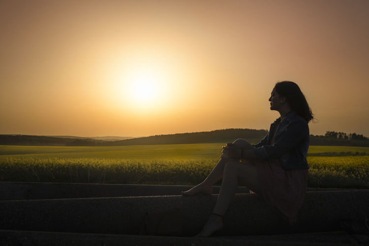 Side view of thoughtful woman looking at view while sitting on field against sky during sunset