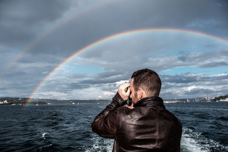 Scenic view of man taking photograph of  rainbow in front of sea against sky