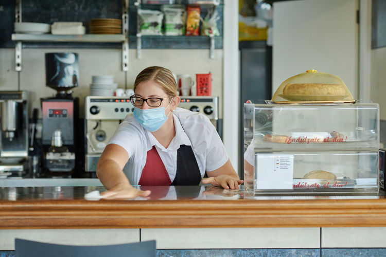 Female bartender in medical mask wiping counter with antibacterial nap while working in cafeteria during coronavirus pandemic