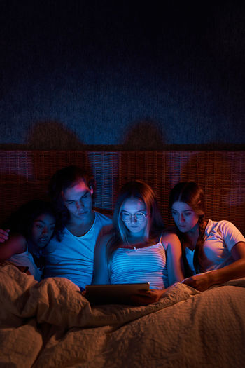 Frightened young friends in sleepwear watching horror on tablet while lying together on comfy bed at night