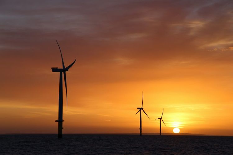 Silhouette wind turbine by sea against sky during sunset