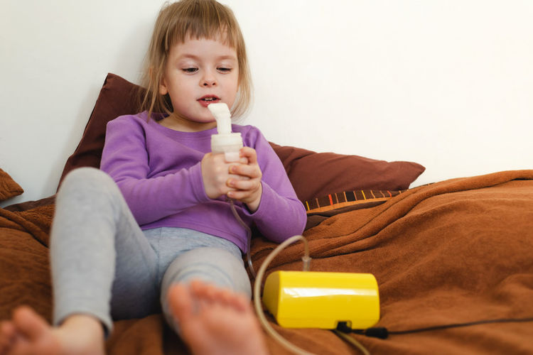 Sick child lies in bed and makes inhalation using a nebulizer.the girl holds an inhaler in her hands