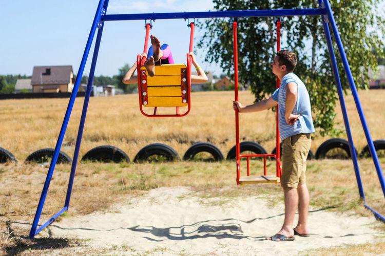 Little girl swinging on swing on playground with young man, guy, her older brother. countryside 