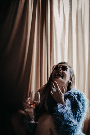 Young woman in sunglasses with glass of wine, blue fur coat and silver boho jewelry laughing