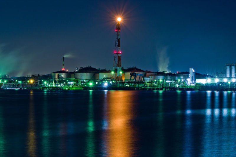 Illuminated factory buildings by sea against sky at night