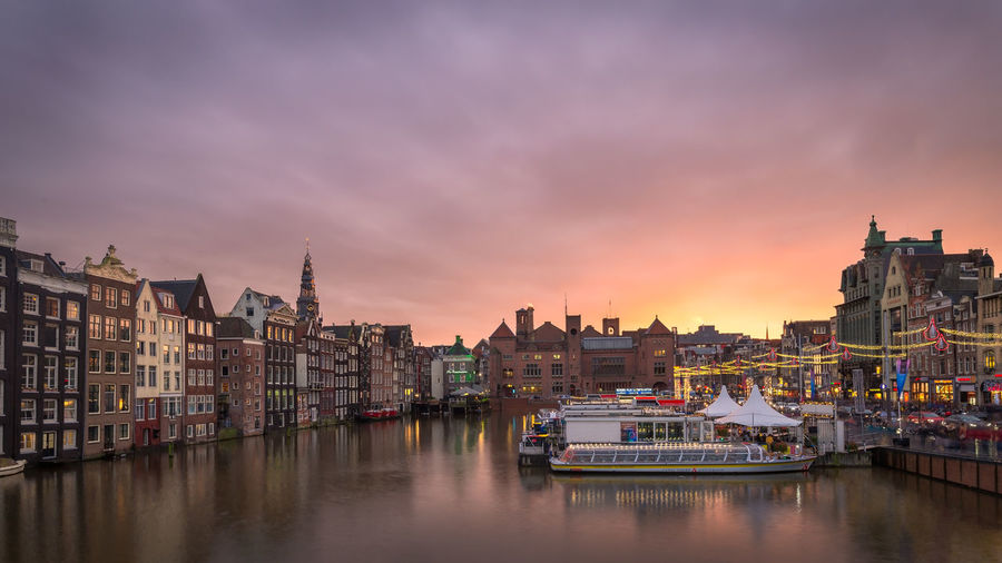 Boats moored in river by buildings against sky during sunset