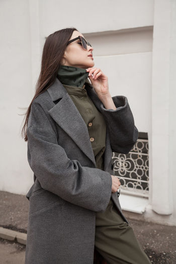 Close up side view postttait of young woman in grey coat standing against wall outdoors, outwear 