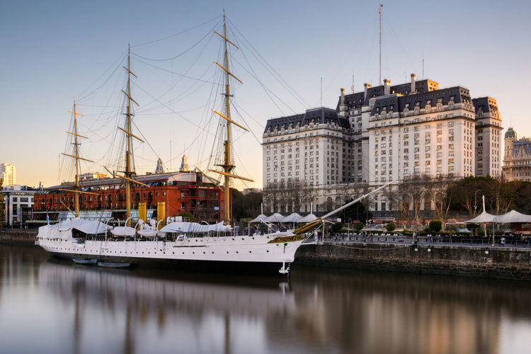 Sailing ship moored in river by buildings against clear sky