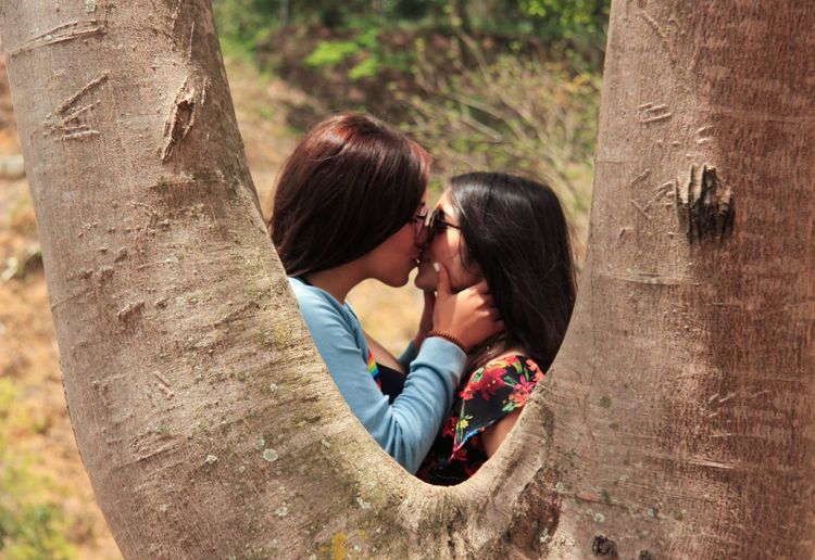 Side view of lesbian couple kissing on mouth seen through tree trunk at public park