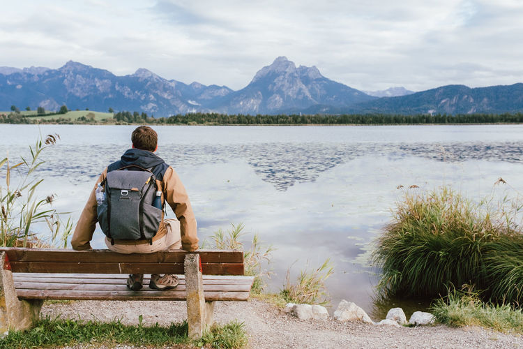 Rear view of man sitting on bench by lake against mountains
