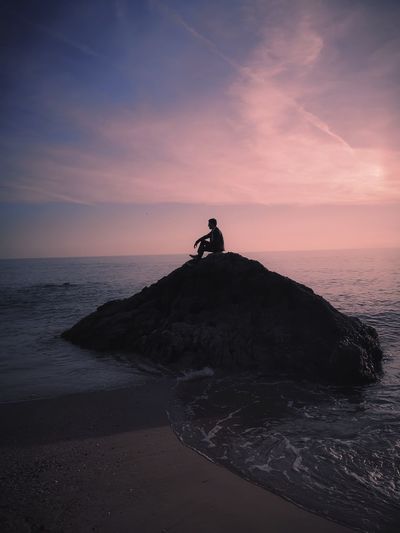 Man on rock by sea against sky during sunset