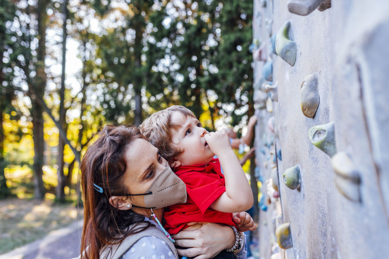 Side view of mother wearing mask holding son by climbing wall