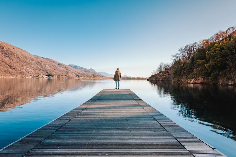 Rear view of man walking on pier over lake against clear blue sky