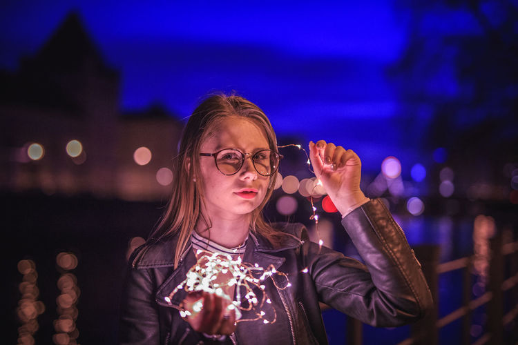 Portrait of young woman holding illuminated string light by lake at night