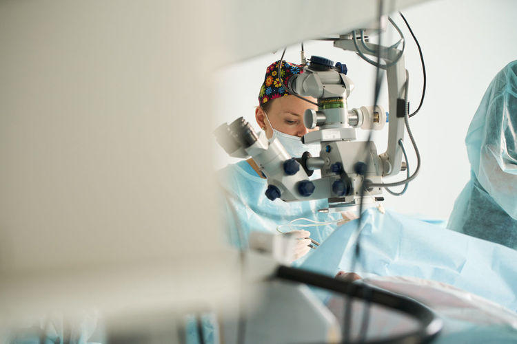 Attentive female doctor in surgical uniform and sterile mask looking through microscope while operating eye of unrecognizable patient in hospital