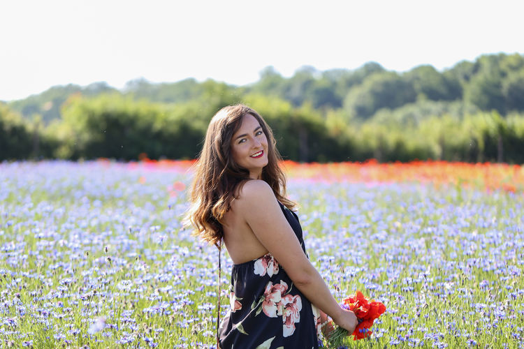 Portrait of smiling young woman holding flowers while standing by plants against sky