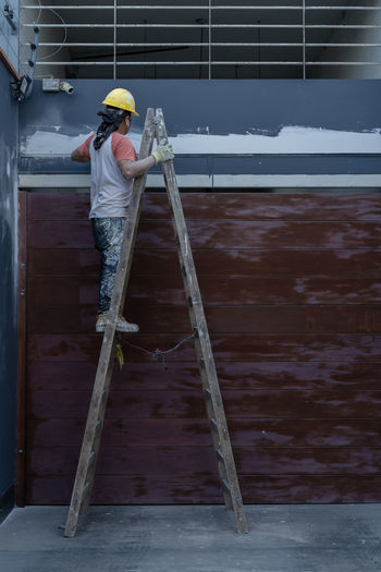 Side view of professional male painter in yellow hardhat in process of plastering wall while standing on a ladder on sidewalk on city street