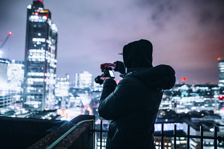 Side view of man photographing illuminated cityscape against sky at night