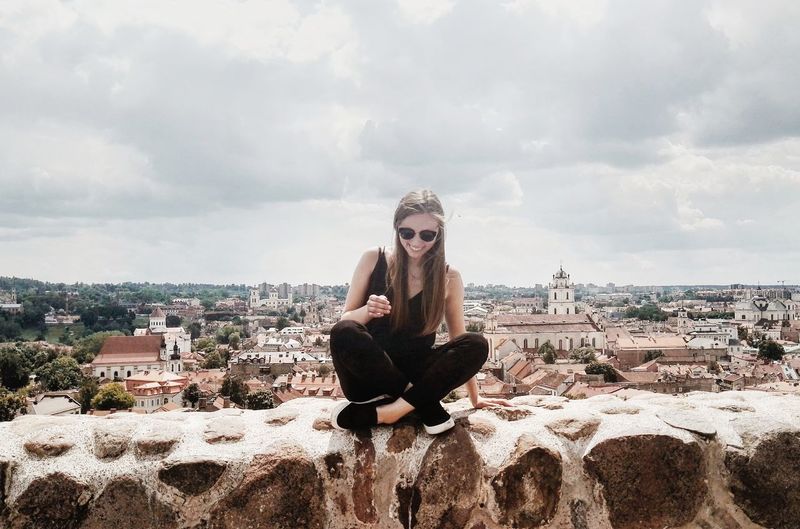 Young woman sitting on retaining wall at vilnius old town against sky
