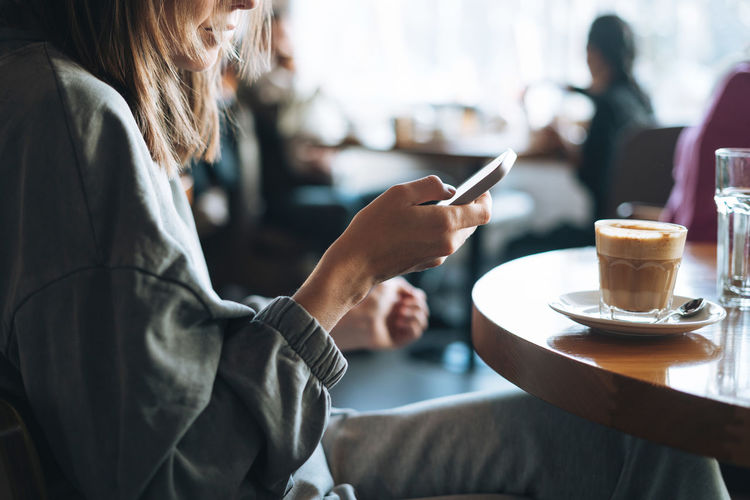 Young brunette woman wearing casual longsleeve with mobile phone in hands with cup of coffee in cafe
