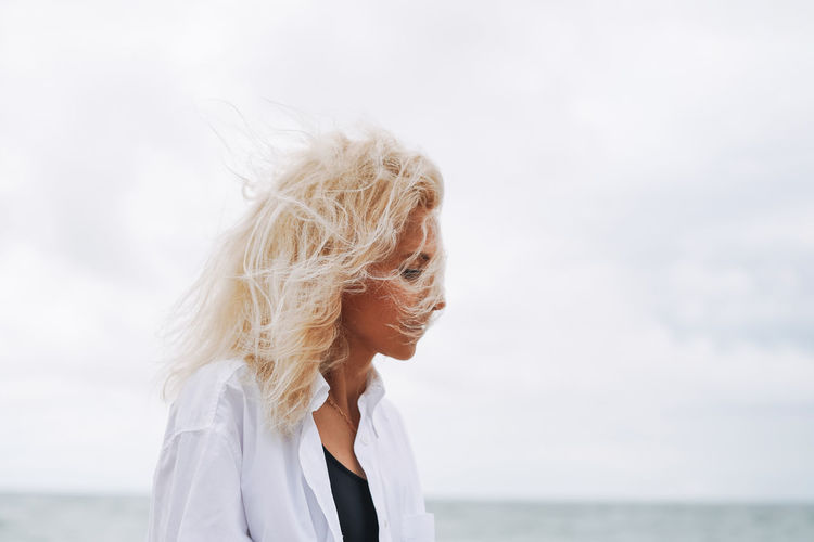 Portrait of elegant blonde woman in white shirt on sand beach at storm sea at windy weater