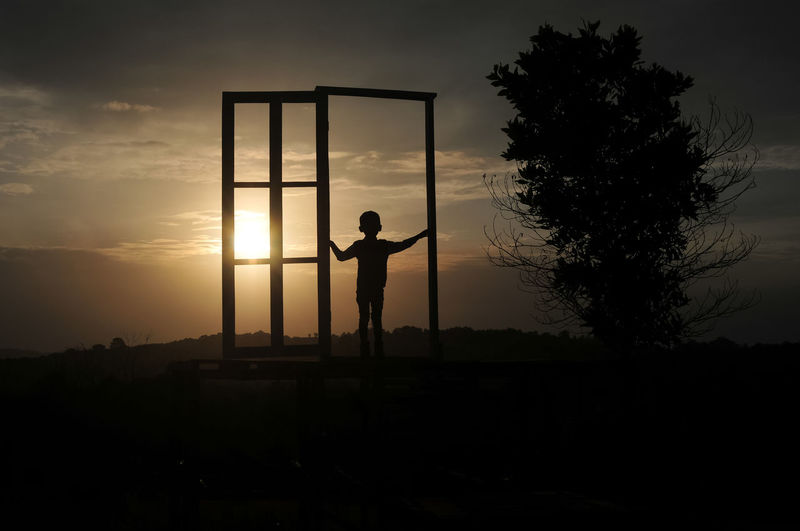 Silhouette boy standing at gate on field against sky during sunset
