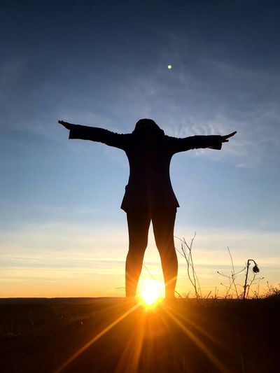 Rear view of silhouette woman with arms outstretched standing on land against sky during sunset